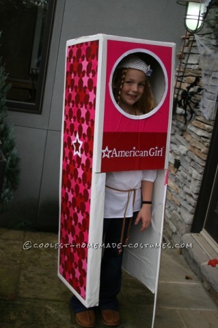 americangirl A girl is standing inside a pink and white box with her head showing through a cut-out. The American Girl logo is on the box.