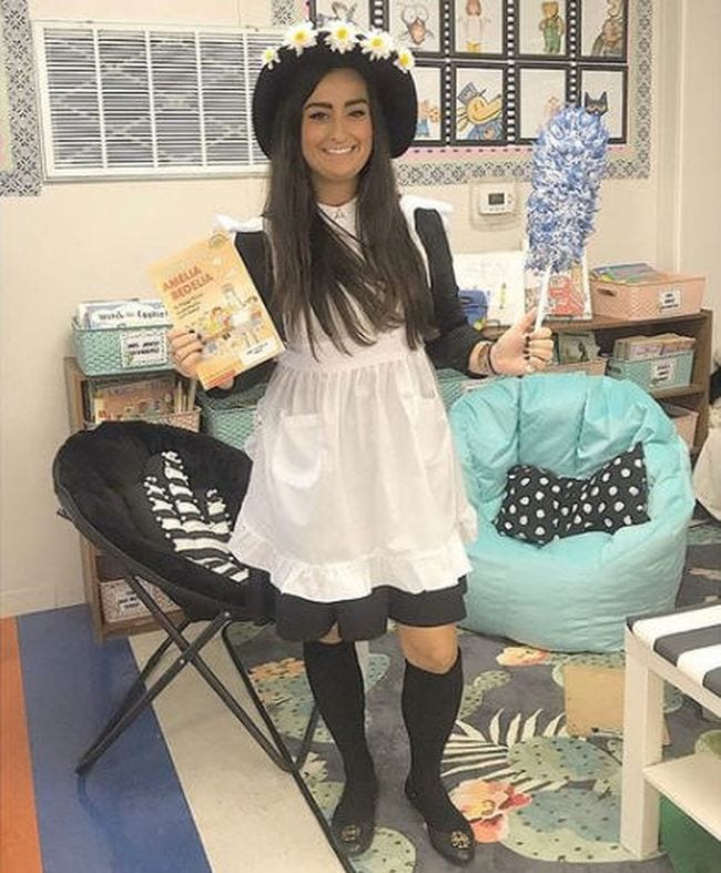 Book Character Costume Ideas Amelia Bedelia Woman wearing black dress, white apron, black hat with daisies, and carrying a feather duster (Book Character Costume Ideas)