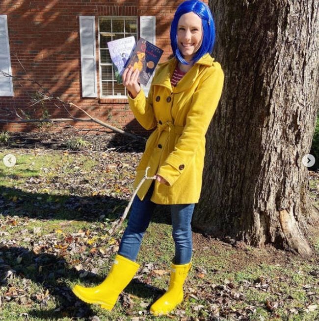 Book Character Costume Ideas Coraline A woman stands in yellow rainboots, a yellow trenchcoat, and a blue wig, holding a key necklace.