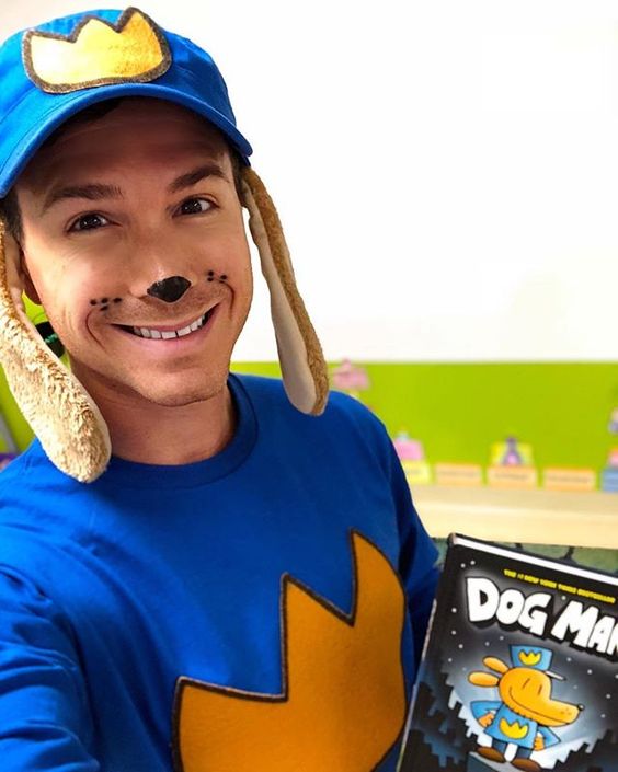 Dog Man A teacher wears a blue hat with a yellow crown on it and dog ears as well as a blue sweatshirt with a yellow crown on it. The teacher's nose is painted black and he's holding the book Dog Man.- book character costume