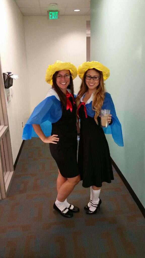 Madeline Two women are seen wearing matching outfits. They are wearing yellow hats, navy dresses, red neckerchiefs, and blue capes.