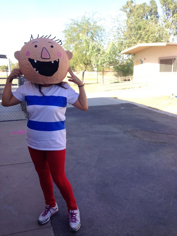 A teacher has on red pants, a white and blue stripe shirt, and a mask with a face on it that is supposed to look like the David character from the book.