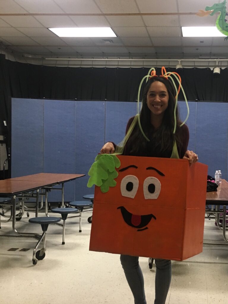 A woman is seen wearing an orange box around her that has a face on it and is meant to look like a pumpkin.