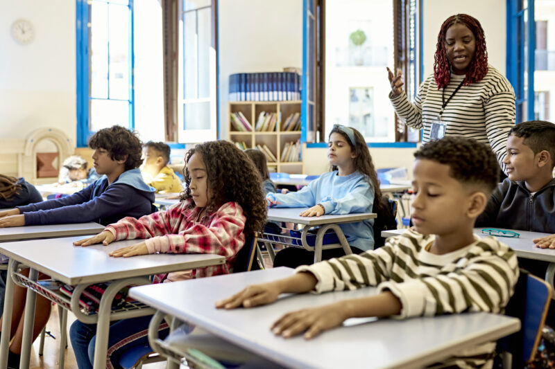 Mature Female Teacher Directing Students During Quiet Time Front view of elementary age schoolchildren sitting at their child-size desks with arms relaxed and eyes closed, enjoying a break from studies.