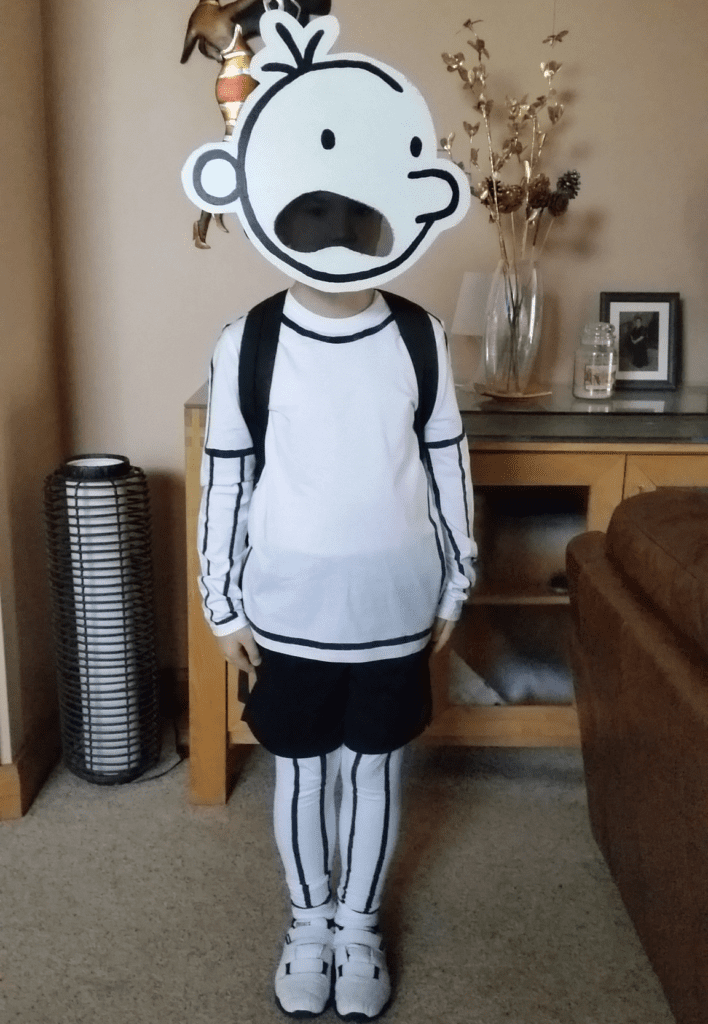 Diary of a Wimpy Kid A person wearing a white shirt, black shorts, and white leggings is standing. They also have a cut out of a simply drawn face like the one the character in Diary of a Wimpy Kid has.